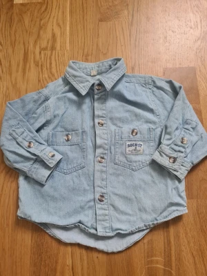 Ljusblå jeansskjorta från Rock-It - Ljusblå jeansskjorta från Rock-It i storlek 86 med klassisk krage, två bröstfickor och knappar framtill. Skjortan har långa ärmar och en avslappnad passform. Perfekt för en cool och avslappnad stil.