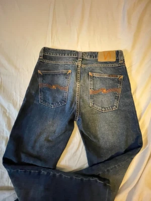 Blå jeans från Nudie Jeans, W27 L30 - Säljer ett par klassiska blå jeans från Nudie Jeans med snygga kontrastsömmar och ikonisk broderad detalj på bakfickorna. Jeansen har normal passform och är tillverkade i bomull. Perfekta för dig som gillar stilrena och tidlösa jeans.