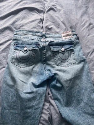 Ljusblå True Religion jeans - Säljer ett par ljusblå jeans från True Religion med raka ben och slitningar vid knäna. Jeansen har klassiska bakfickor med lock och vita knappar samt dekorativa sömmar. Materialet är denim i bomull och passformen är relaxed.