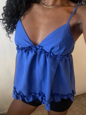 Blå linnetopp med volangdetaljer - Snygg blå linnetopp med tunna axelband och djup v-ringning. Toppen har volangdetaljer både under bysten och längs nederkanten för en extra feminin touch. Perfekt för varma dagar och enkel att styla till olika looks.