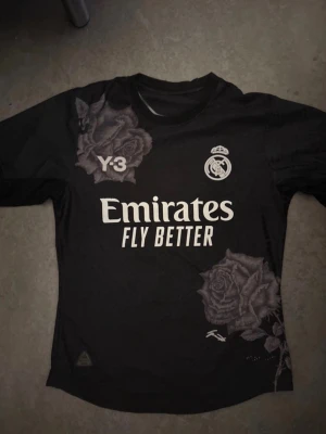 Real Madrid x Y-3 svart matchtröja - Snygg svart Real Madrid matchtröja i samarbete med Y-3 och Adidas. Tröjan har vita tryck med klubbmärke, sponsorlogga och coola rosdetaljer både fram och bak. Tillverkad i lätt och funktionellt material, perfekt för träning eller match.
