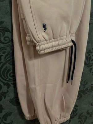 Beiga mjukisbyxor från Polo med marinblå snören - Snygga beiga mjukisbyxor från U.S. Polo Assn. med marinblå dragsnören och broderad logga på benet. Byxorna har resår i midjan och vid bensluten för en skön och avslappnad passform. Perfekta för chill dagar eller när du vill vara bekväm.