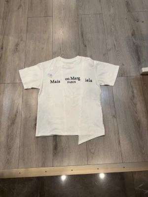 Vit Maison Margiela t-shirt med asymmetrisk kant - Vit t-shirt från Maison Margiela med svart tryck på bröstet och unik asymmetrisk nederkant. Klassisk rund halsringning och korta ärmar. Tillverkad i mjuk bomull för en skön känsla och modern look.