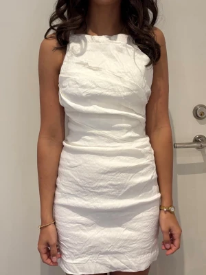 Vit ärmlös kort klänning - Säljer en stilren vit kort klänning med hög hals och figurnära passform. Klänningen är ärmlös och har en enkel, elegant design som passar perfekt till sommarens alla tillfällen. Materialet känns lätt och bekvämt. Perfekt till studenten!
