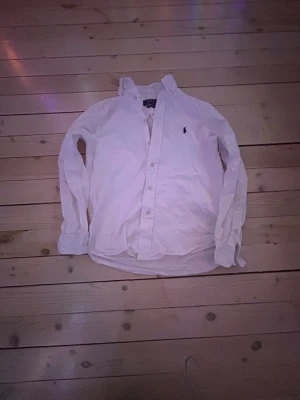 Vit skjorta från Ralph Lauren - Klassisk vit skjorta från Ralph Lauren med den ikoniska broderade loggan på bröstet. Skjortan har lång ärm, knappar framtill och en tidlös passform. Perfekt till jeans eller chinos för en clean och stilren look.säljer denna skjorta pga av att den har blivit för liten.nypris runt 1000kr