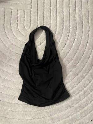 Svart halterneck topp - Säljer en svart halterneck topp med draperad urringning. Toppen är ärmlös och har en tight passform som framhäver figuren. Perfekt att styla med jeans eller kjol för en trendig look.