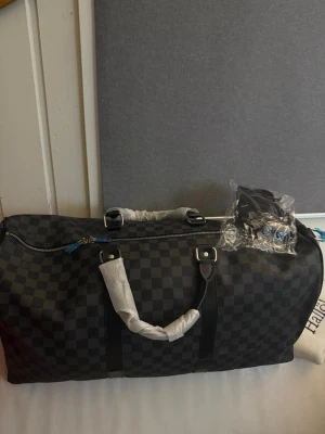 Louis Vuitton keepall  - Snygg lv weekendbag med klassiskt rutmönster i svart och grått. Väskan har dubbla handtag, silverfärgade metalldetaljer och dragkedja. Perfekt för resor eller gymmet. Materialet är slitstarkt och handtagen är förstärkta för extra hållbarhet.