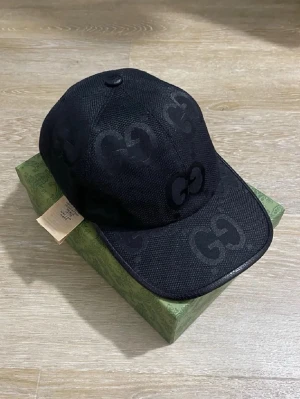 Svart keps från Gucci med GG-mönster - Snygg svart keps från Gucci med klassiskt GG-monogram i ton-i-ton. Kepsen har böjd skärm, justerbar rem baktill och är tillverkad i ett slitstarkt tyg med mesh-känsla. Perfekt accessoar för att lyfta din streetwear-look.