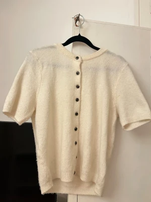 Krämvit kofta från H&M med knappar - Mysig krämvit kofta från H&M med korta ärmar och svarta knappar framtill. Koftan har rund halsringning och är stickad i ett mjukt material som känns fluffigt. Perfekt att slänga över en topp för en chill look.