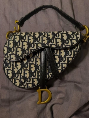  handväska med monogram - Snygg väska med ikoniskt svart och beige monogrammönster. Väskan har en unik sadelform, guldfärgade metalldetaljer. Detta är inte den stora modellen utan den mindre, den är perfekt för utgångar/middagar.