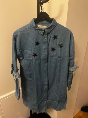 Oversized jeansskjorta med stjärnor - Säljer en oversized jeansskjorta från Zara i blå denim med svarta glittriga stjärnapplikationer på bröstet. Skjortan har långa ärmar med knytband vid ärmsluten och klassisk krage. Perfekt för en avslappnad och trendig look.