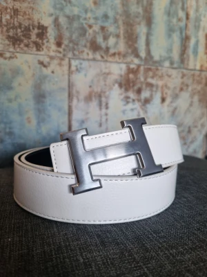 Vitt skärp med H-spänne - Snyggt vitt skärp med ett stort, silverfärgat H-spänne. Skärpet är gjort i slätt skinn och har stilrena sömmar längs kanterna. Perfekt accessoar för att lyfta vilken outfit som helst.