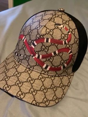 Gucci keps med orm och GG-mönster - Säljer en snygg keps från Gucci med klassiskt GG-monogram i beige och brunt samt en röd och vit orm på framsidan. Kepsen har svart mesh baktill och justerbar rem. Perfekt för dig som vill sticka ut med en lyxig accessoar. För mer bilder är det bara och skriva!