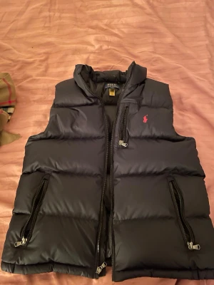 Svart dunväst från Polo Ralph Lauren - Svart dunväst från Polo Ralph Lauren med två dragkedjefickor och klassisk röd logga på bröstet. Västen har hög krage och breda kanaler för en puffig look. Perfekt för lager-på-lager och streetstyle.