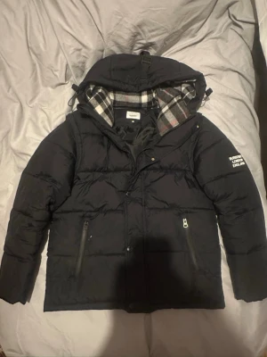 Svart pufferjacka från Burberry - Snygg svart pufferjacka från Burberry med avtagbar huva och klassiskt rutigt foder i grått, vitt och rött. Jackan har två dragkedjefickor fram och tryckt text på ärmen. Perfekt för kalla dagar och riktigt trendig look.