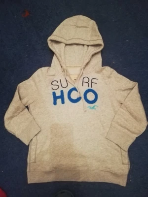 Beige hoodie från Hollister med tryck - Snygg beige hoodie från Hollister med blått och svart tryck 'SURF HCO' på bröstet. Klassisk huva och känguruficka framtill. Tillverkad i mjukt bomullsmaterial, perfekt för chill dagar eller när du vill ha en avslappnad look.