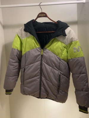 Grå och limegrön pufferjacka med huva - Säljer en riktigt fet pufferjacka i grått, limegrönt och vitt. Jackan har en stor huva, dragkedja framtill och två sidofickor med dragkedja. Snygg detalj med ett tryckt märke på ärmen. Perfekt för kalla vinterdagar och riktigt skön att bära.