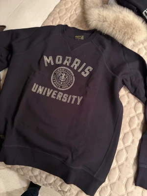 Svart sweatshirt från Morris - Collegeinspirerad svart sweatshirt från Morris med trycket 'Morris University' och emblem på bröstet. Tröjan har rund halsringning, ribbade muddar och är tillverkad i mjukt bomullsmaterial. Perfekt för en avslappnad och stilren look.