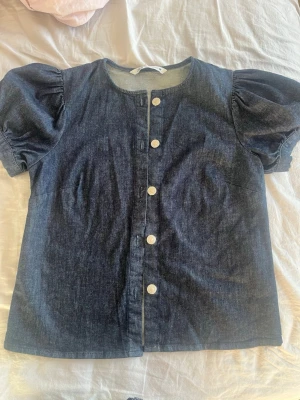 Mörkblå jeansblus med puffärm - Snygg mörkblå jeansblus med puffiga korta ärmar och vita knappar framtill. Rak passform och rund halsringning. Tillverkad i denimtyg som ger en cool och trendig look.