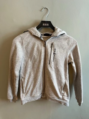 Ljusgrå hoodie från Sail Racing - Ljusgrå hoodie från Sail Racing med dragkedja framtill och en bröstficka med logga. Tröjan har två sidofickor och en skön huva med tryckt detalj bak. Tillverkad i mjukt sweatshirtmaterial, perfekt för chill dagar. Pris kan anpassas