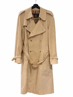 Vintage Burberry Trench Coat - Vintage Burberrys trenchcoat – Made in England  Klassisk beige trenchcoat från Burberrys (vintage, äldre logga).  Tillverkad i England med ikoniskt rutigt foder.  Bra vintage-skick med normalt bruksslitage, har ett litet märke/skada vid vänster fram-axel – se inzoomad bild.  Uppskattad storlek: L Bröstmått: cirka 106 cm Längd: cirka 111 cm  Hör av dig om du har frågor eller vill ha fler bilder!