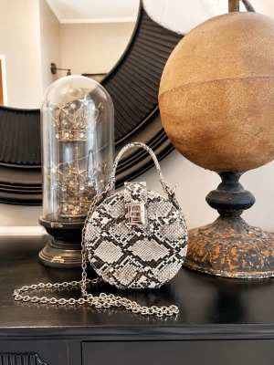 Rund handväska i ormmönster - Säljer en rund handväska med svartvitt ormmönster och silverfärgad kedjerem. Väskan har ett metallspänne framtill och är tillverkad i konstläder. Perfekt för dig som vill sticka ut med en unik accessoar.