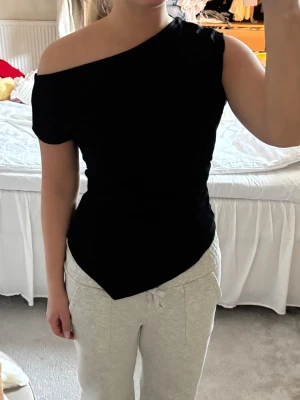 Svart oneshoulder topp - Snygg svart topp med oneshoulder-design och asymmetrisk nederkant. Toppen är tight och har kort ärm på ena sidan medan andra axeln är bar. Perfekt för dig som vill ha en trendig och stilren look! Har inte kommit till användning så därav säljer jag den🖤
