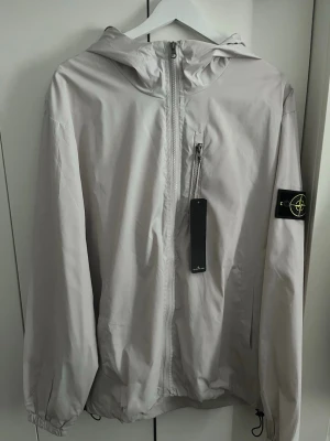 Stine Island Windbreaker - Snygg ljusgrå vindjacka från Stone Island med huva och dragkedja framtill. Jackan har en klassisk patch med logga på vänster ärm och justerbar nederkant. Tillverkad i lätt, vindtätt material som passar perfekt till streetwear-stilen.
