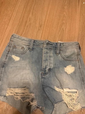 Ljusblåa jeansshorts hollister - Säljer ett par ljusblåa jeansshorts med slitna detaljer och fransig kant. Shortsen har klassisk femficksmodell, bälteshällor och dragkedjegylf. Perfekta för sommaren och ger en avslappnad vibe.