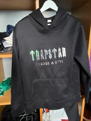 Svart Trapstar hoodie med tryck - Säljer en svart hoodie från Trapstar med stort tryck i grönt och vitt på bröstet där det står 'Trapstar It’s a secret'. Hoodien har huva och en klassisk magficka. Perfekt för dig som gillar streetwear och vill sticka ut med en unik design.