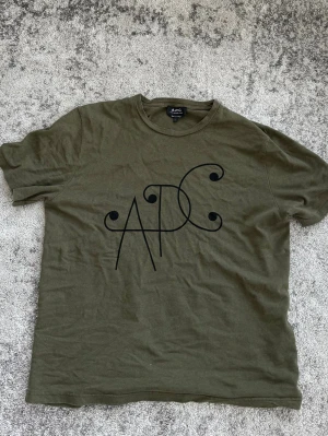 Olivgrön APC t-shirt med tryck - Snygg olivgrön t-shirt från A.P.C. med ett stort svart grafiskt tryck på bröstet. Klassisk rund halsringning och korta ärmar. Perfekt för en avslappnad och stilren look. Perfekt skick. Storlek L men passar även M