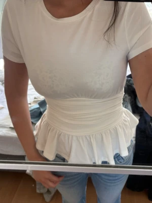 Vit peplum topp med markerad midja - Supersöt vit topp med korta ärmar och peplumvolang nedtill. Sitter tajt över midjan och har en rund halsringning. Perfekt att styla med jeans för en trendig look.