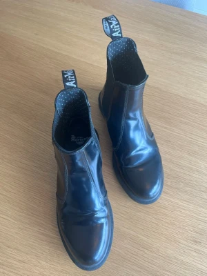 Svarta Chelsea boots Dr. Martens - Säljer ett par klassiska svarta Chelsea boots från Dr. Martens, modell Flora. Skorna är i blankt läder med elastiska paneler på sidorna och har den ikoniska Dr. Martens-flärpen bak. Perfekta för dig som vill ha en stilren och edgy look.