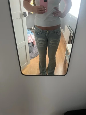 Ljusblå bootcut jeans med slitningar - Säljer ett par ljusblå bootcut jeans med snygga slitningar och tvättade detaljer över låren. Jeansen har låg midja, klassiska fem fickor och bälteshällor. Perfekta för en avslappnad och trendig look.