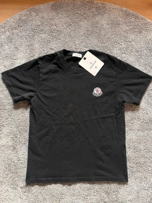 Svart Moncler t-shirt med logga - Snygg svart t-shirt från Moncler med klassisk rund hals och korta ärmar. På bröstet sitter Monclers ikoniska broderade logga i vitt, blått och rött. Tillverkad i mjuk bomull för en skön känsla och enkel stil.