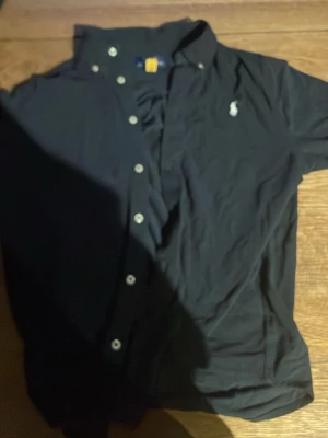 Mörkblå skjorta från Ralph Lauren - Klassisk mörkblå skjorta från Ralph Lauren med vit broderad logga på bröstet. Skjortan har knappar hela vägen, button-down-krage och lång ärm. Perfekt till jeans eller chinos för en clean och stilren look.