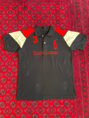Svart pikétröja från Ralph Lauren - Svart pikétröja från Ralph Lauren med röda detaljer, stor röd logga och text på bröstet samt siffran 3 på axeln. Tröjan har korta ärmar med vita och röda partier upptill och klassisk krage med knappar. Perfekt för en sportig och stilren look.