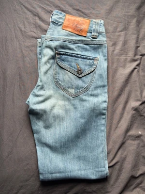 Blå jeans från Lindex med fickdetaljer - Snygga blå jeans från Lindex med ljus tvätt och coola fickdetaljer både fram och bak. Jeansen har en klassisk femficksmodell, knappgylf och bälteshällor. Materialet är mjukt denim i bomull med lite stretch för extra komfort.