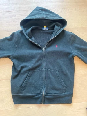 Svart hoodie från Polo Ralph Lauren - Klassisk svart hoodie från Polo Ralph Lauren med dragkedja, huva och kängurufickor. Ikoniska röda loggan broderad på bröstet. Tillverkad i mjukt bomullsmaterial som är skönt mot huden. Perfekt för en avslappnad och stilren look.