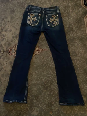 Blå bootcut jeans med broderi - Säljer ett par blå bootcut jeans från Miss Me med vita kontrastsömmar och snygga broderade detaljer på bakfickorna. Jeansen har låg midja, fem fickor och slitningar framtill för en cool look. Perfekta för dig som gillar unik stil!