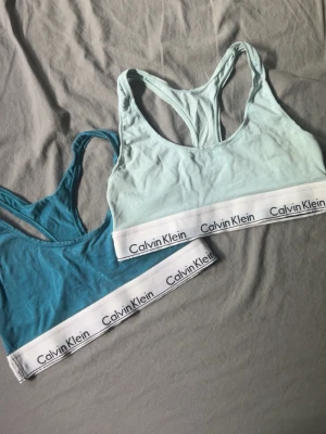 Två Calvin Klein bh-ar  - Två bhar från Calvin Klein i ljusblått och turkost. Båda har bred resår med logga, brottarrygg och är tillverkade i mjukt bomullsmaterial. 