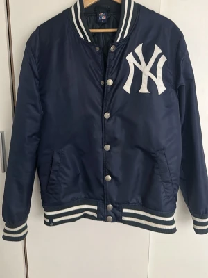 New York Yankees Varsity Jacket Navy Size L - Säljer en New York Yankees varsity jacket i marinblått.  Klassisk baseballjacka med stort Yankees-logo framtill. Perfekt för en clean streetwear-look. Bra skick.  Storlek: L men passar M Färg: Marinblå/Vit  Skickar gärna fler bilder vid intresse.