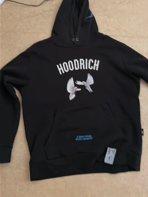 Svart hoodie från Hoodrich med duvor - Svart hoodie från Hoodrich med tryck av vita duvor och texten 'HOODRICH' på bröstet. På ryggen finns fler duvor och texten 'From Nothing To Something'. Hoodien har huva och magficka, samt detaljer i blått och vitt. Perfekt för en streetwear-look.