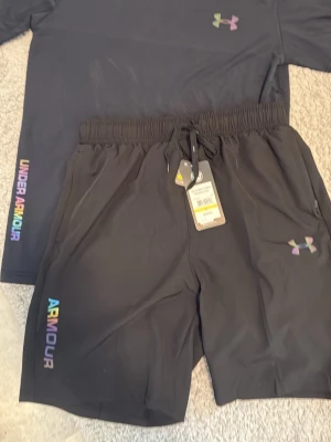 Svarta shorts från Under Armour - Snygga svarta shorts från Under Armour med elastisk midja och snörning. Loggan och texten 'ARMOUR' i regnbågsfärger ger en cool touch. Perfekta för träning eller chill. Tillverkade i ett lätt och snabbtorkande material.