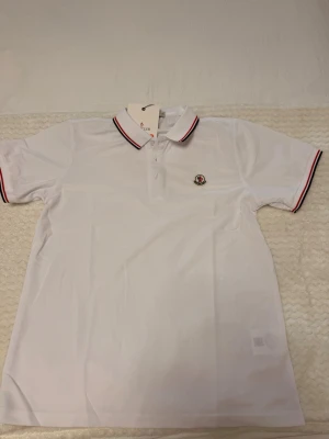 Vit Moncler pikétröja med röd detalj - Säljer en riktigt snygg Moncler pikétröja i vit färg, en tidlös och exklusiv modell som ger en fräsch och stilren look. Den eleganta vita färgen tillsammans med de klassiska röd/vita detaljerna på kragen och ärmarna ger ett lyxigt intryck och gör tröjan enkel att matcha till både jeans, shorts eller mer uppklädda outfits.