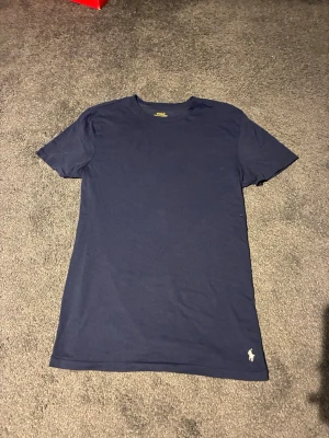Mörkblå t-shirt från Ralph Lauren - En stilren mörkblå t-shirt från Ralph Lauren med klassisk passform och det ikoniska vita Polo-logotypen broderad nertill. Tillverkad i mjuk bomull och har korta ärmar. Perfekt till jeans eller shorts för en clean look.