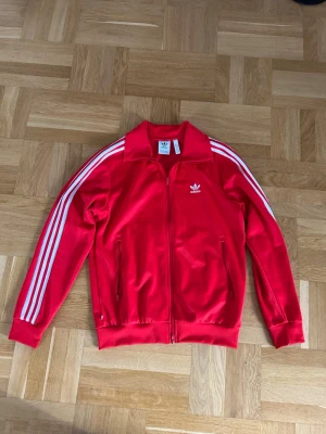 Röd Adidas track jacket med vita ränder - Klassisk röd Adidas track jacket med dragkedja, vita ränder längs ärmarna och Adidas-logga på bröstet. Jackan har hög krage, muddar vid ärmslut och nederkant samt två fickor med dragkedja. Tillverkad i ett glansigt syntetmaterial.