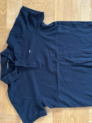 Svart piké från J. Lindeberg - Snygg svart piké från J. Lindeberg med klassisk krage och två knappar framtill. T-shirten har korta ärmar och är tillverkad i mjuk bomull, perfekt för en avslappnad och stilren look.