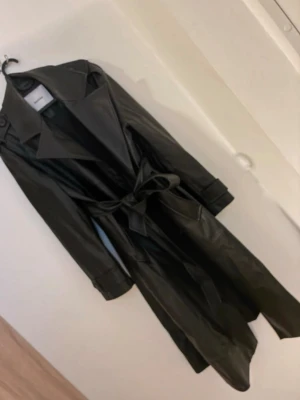 Svart trenchcoat  Bershka - Hål i ena ficka inget som syns på utsidan lite smink fläck vid krage men går nog att tvätta bort. Stilren svart trenchcoat från Bershka i fuskläder med klassisk krage, knytskärp i midjan och långa ärmar. .