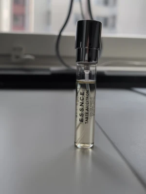 ESSENCE Tarte au Citron parfymprov - Liten parfymflaska från ESSENCE med doften Tarte au Citron. Flaskan är genomskinlig med svart plastlock och innehåller en ljusgul vätska. Perfekt att ha med i väskan för en fräsch citrusdoft.